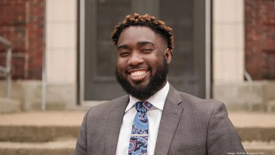 A smiling young man in a gray suit coat and blue tie.
