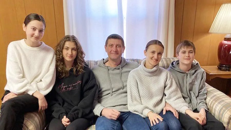 A family of five sits on a couch in front a window.